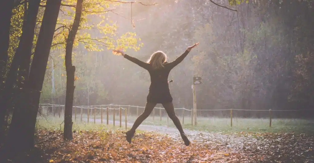 A woman in mid-air, jumping with her arms outstretched in a sunlit forest.