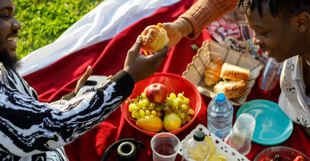 Two people sitting on a red blanket outdoors, sharing food and drinks from a picnic basket.