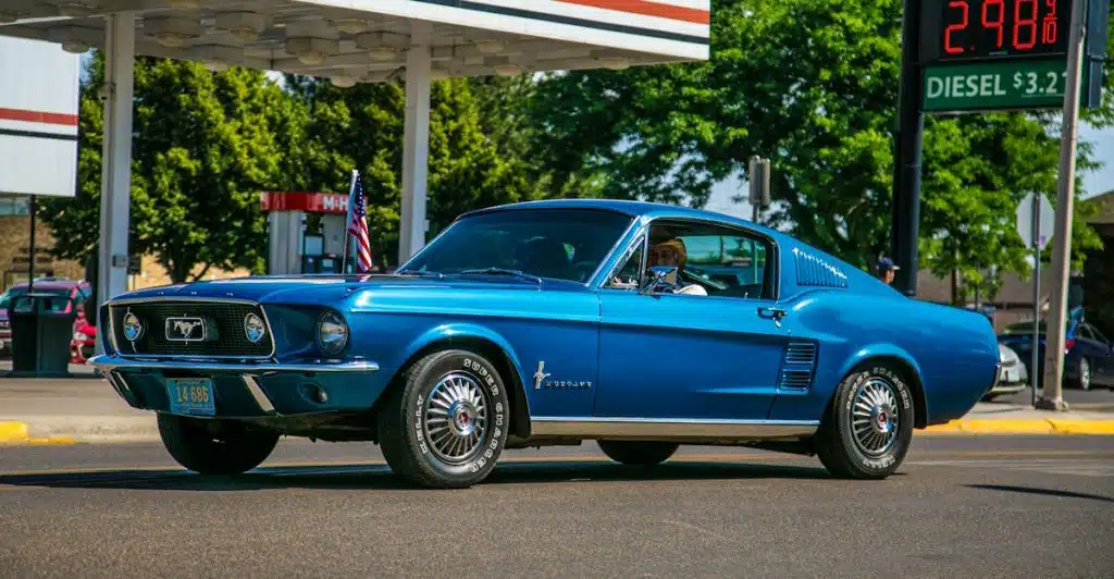 A blue 1967 Ford Mustang Fastback parked at a gas station with a "24 HOURS COLD BEER" sign and gas prices visible.
