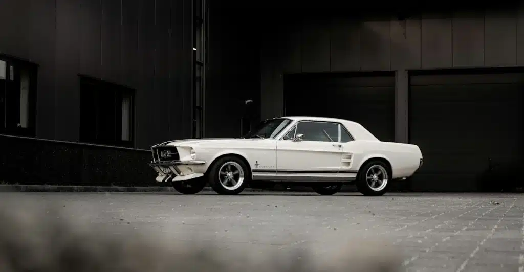 A white classic Ford Mustang with black racing stripes parked on an asphalt lot in front of a dark building.