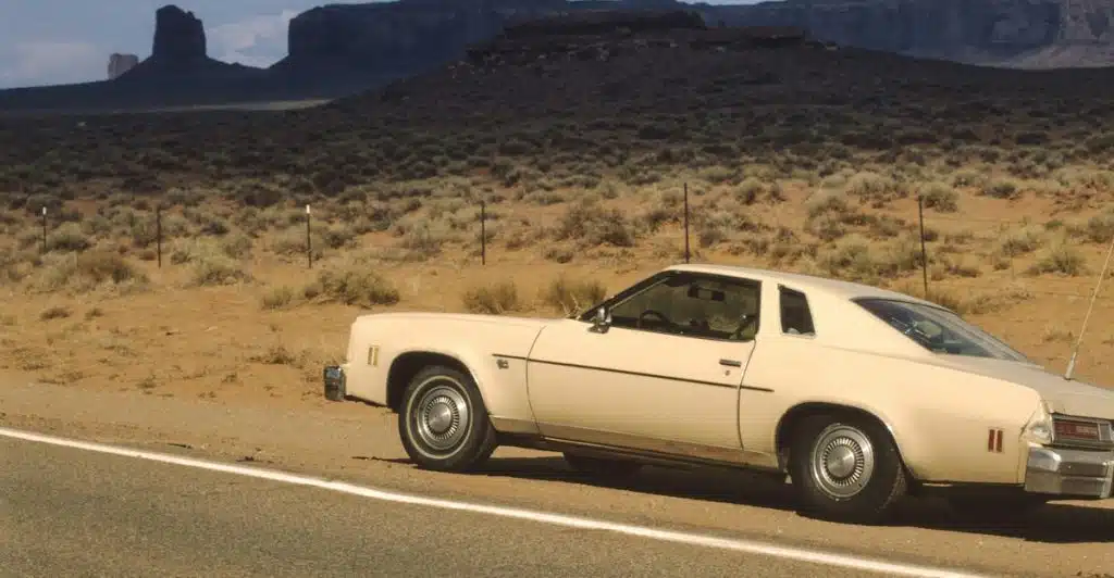 A light-colored classic car parked on the shoulder of a road in a desert landscape with large rock formations in the background.