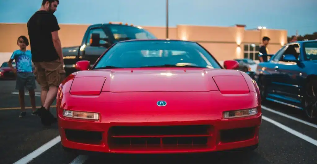 A red 1991 Acura NSX parked at dusk, with people and other vehicles in the blurred background.
