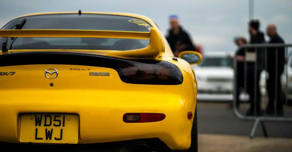 The rear view of a yellow 1992 Mazda RX-7 FD with a large spoiler and a UK license plate, parked outdoors with people in the background.