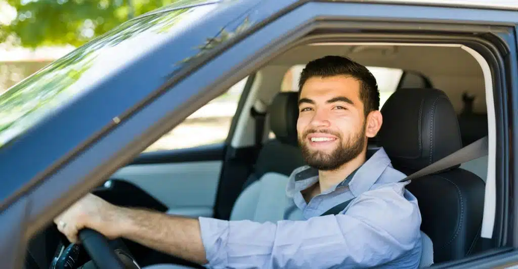 A man with a beard smiling while sitting in the driver's seat of a car, holding the steering wheel.