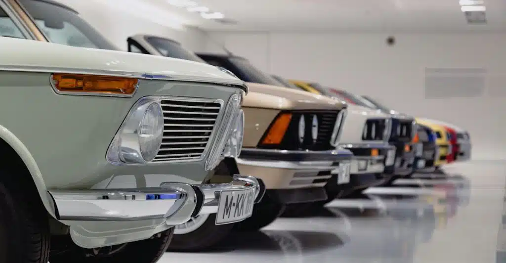 A close-up of the front of several classic cars lined up indoors, showing their headlights and grilles.