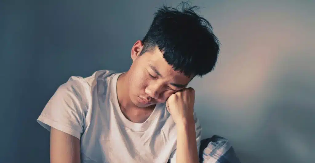 A young man with dark hair asleep at a table, resting his head on his hand.