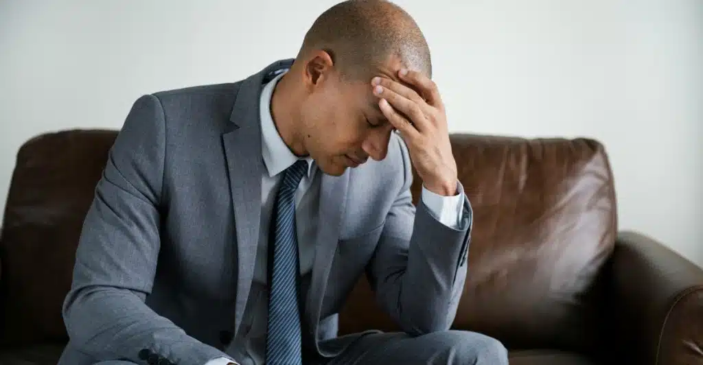 A man in a suit sitting on a couch with his hand on his forehead, appearing stressed.