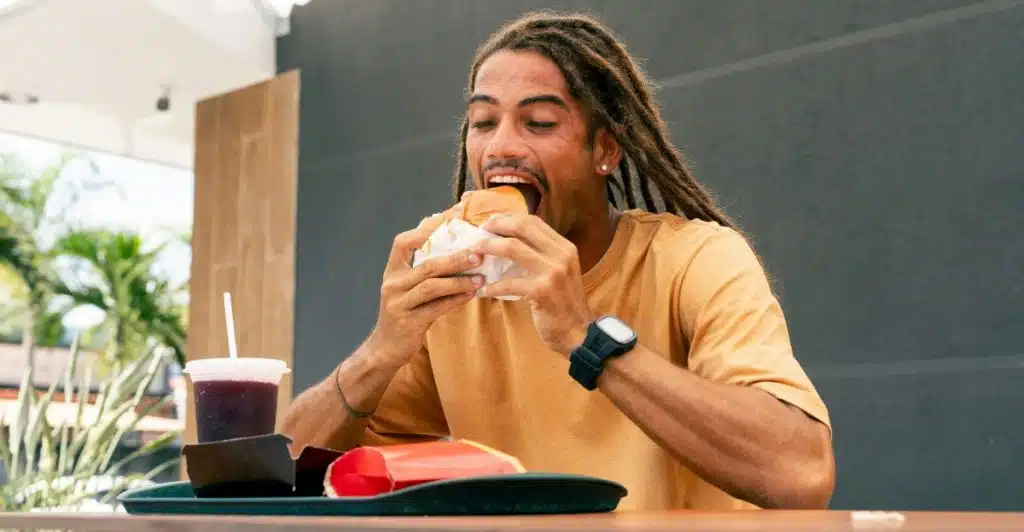 A man with dreadlocks wearing a yellow t-shirt eating a hamburger at an outdoor restauran