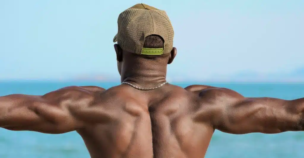 A muscular man from behind, wearing a hat and necklace, standing on a beach with the ocean in the background.