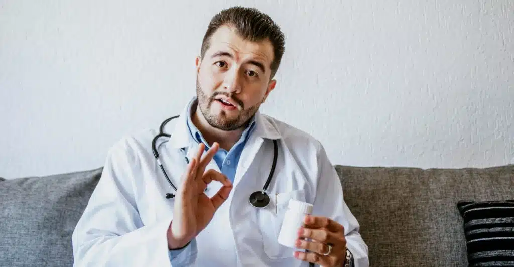 A doctor in a white coat holding a bottle of pills and making an "okay" gesture.