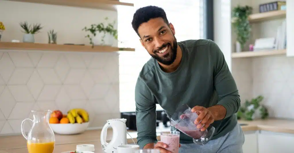 A man in a kitchen smiling and pouring a smoothie into a glass, with fruits and vegetables on the counter.