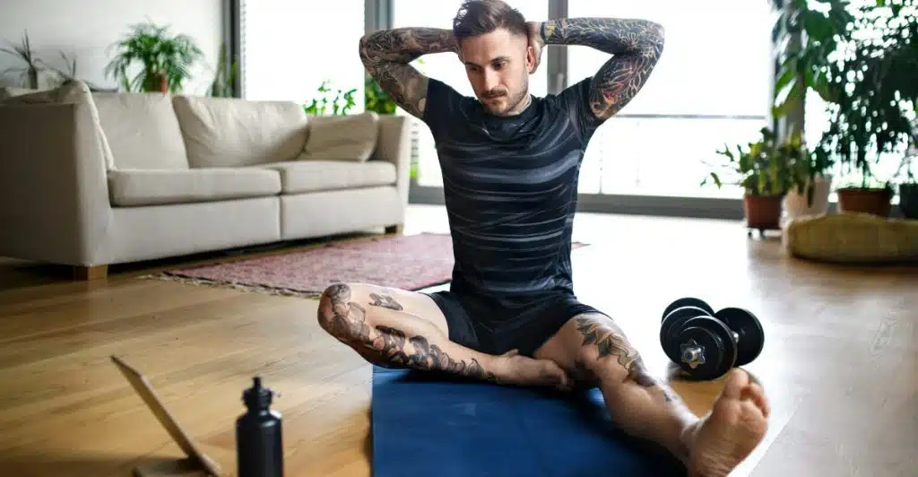 A man with tattoos sitting on a mat at home, stretching his arms behind his head, with dumbbells and a laptop nearby.