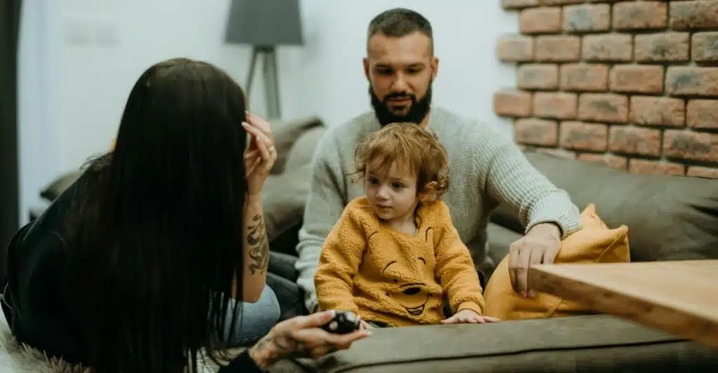 A family of three sitting on a modern sofa in a living room, with a bearded man, a woman with long dark hair, and a small child in a yellow top holding a remote control.