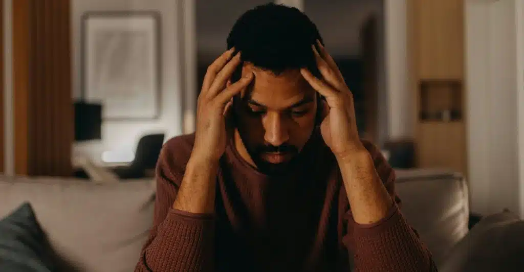 A man with dark skin and a beard sitting indoors in dim lighting, holding his head in his hands with an expression of distress.