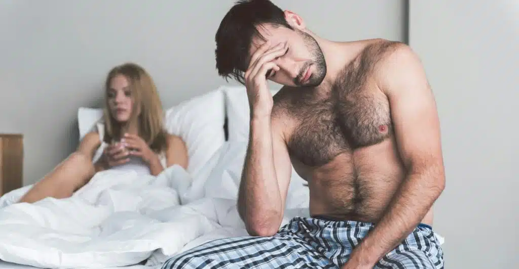 A shirtless man in plaid pajama pants sitting on the side of a bed with his hand covering part of his face in a gesture of frustration, while a woman in a nightgown sits further back on the bed looking away.