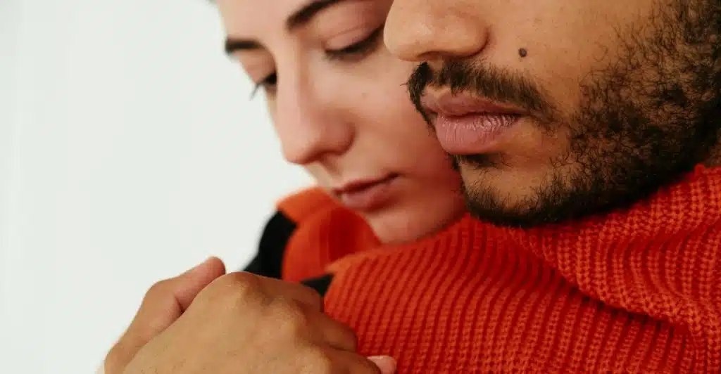 A close-up of a man with a dark beard wearing an orange sweater, tenderly embracing a woman with fair skin and dark hair who is leaning into him.