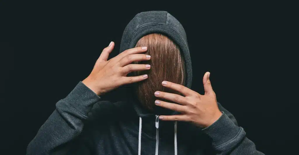 A person wearing a dark hoodie, with their face completely obscured by a mass of long brown hair that they are holding with both hands against a dark background.