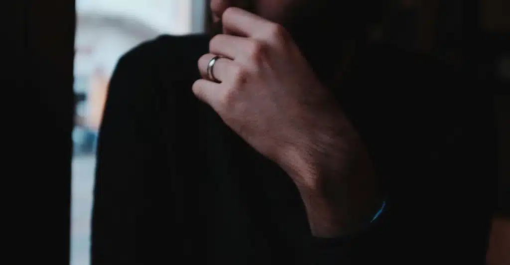 A close-up of a man's left hand, adorned with a wedding ring, raised to his chin as he stands in a dimly lit room, with his face obscured.