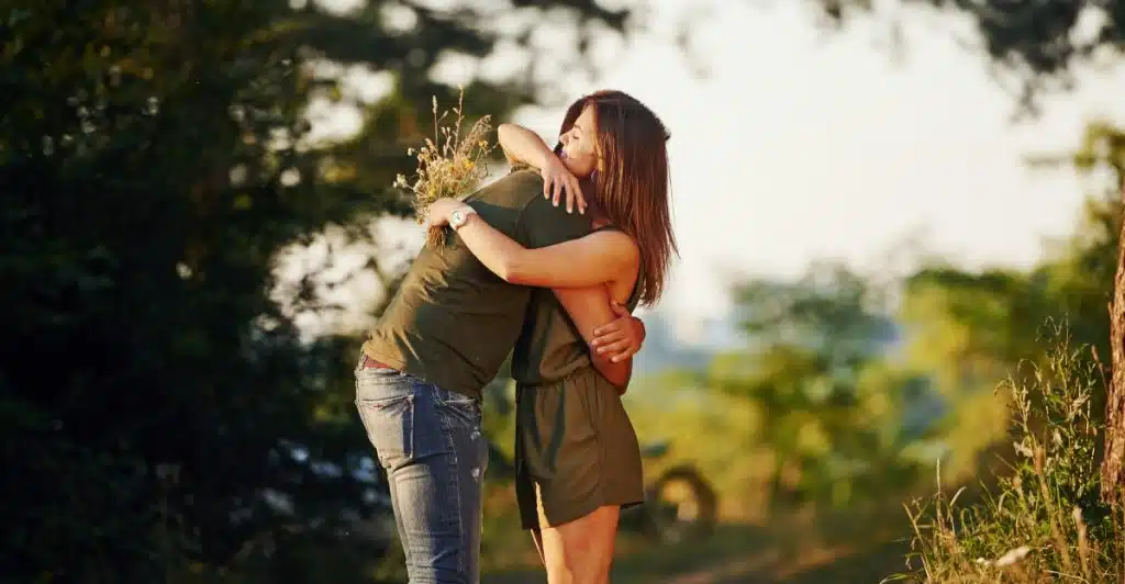 A man and woman embracing in a field with tall grass and trees in the background, with the woman wearing a dress and the man in jeans and a shirt.