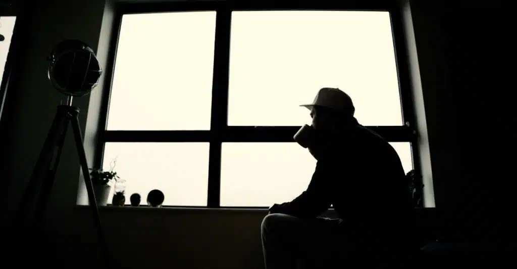 A man in a cap and dark clothing is silhouetted against a large window, holding a cup and looking out, creating a contemplative scene.