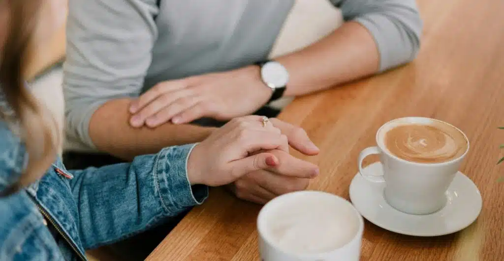A close-up of a man's and a woman's hands intertwined on a light wooden table, with two coffee cups nearby, suggesting a comforting conversation.