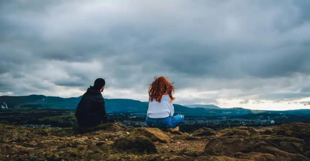 A couple seated side-by-side on a rocky hilltop, looking out at a sprawling landscape under a dramatic, cloudy sky.