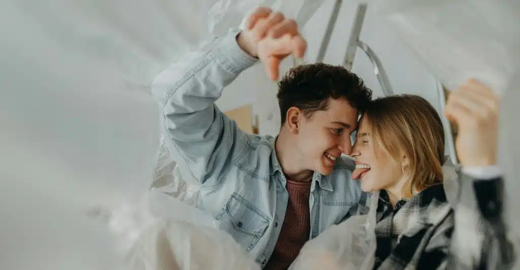 A cheerful couple looking at each other and smiling from underneath clear plastic sheeting, suggesting a playful or intimate moment.