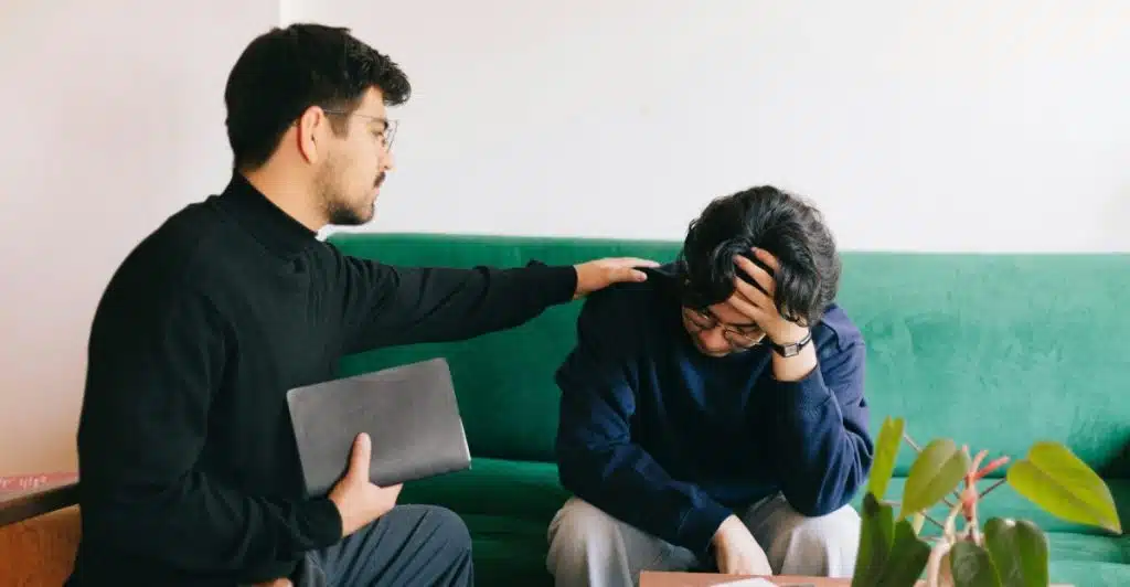 A man with a beard and dark sweater seated next to another man who is distressed with his head in his hands, offering comfort by placing a hand on his shoulder.