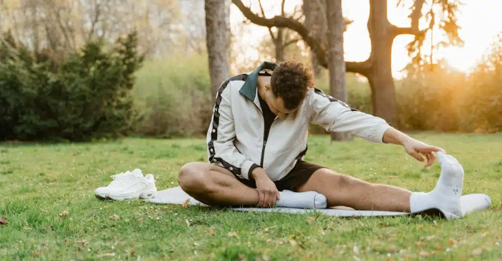 A man with curly hair sitting on a yoga mat in a grassy park, stretching his leg with one hand touching his toes, next to a pair of white sneakers.
