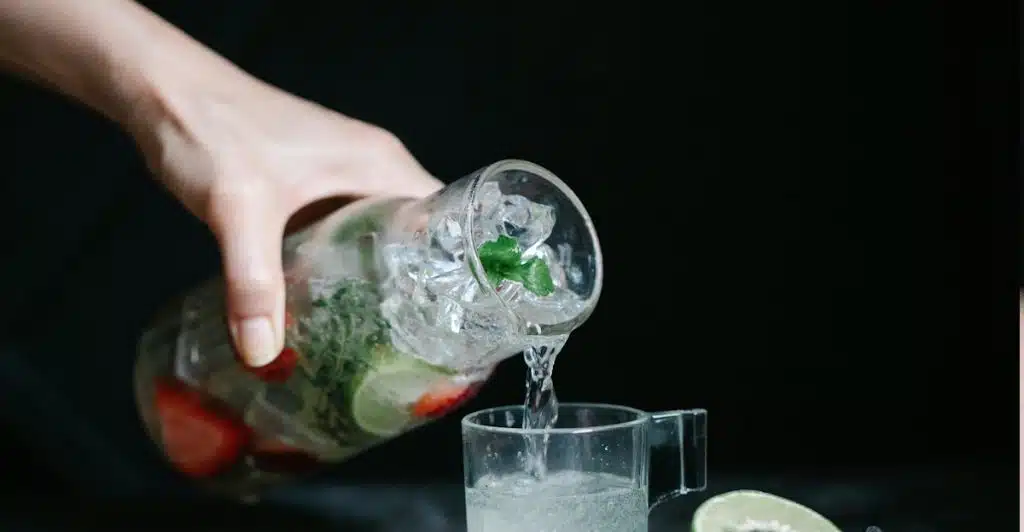 A hand pouring infused water with ice, strawberries, and lime slices from a glass pitcher into a small glass, with additional fruit on a dark surface.