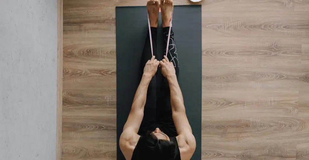 A man lying on his back on a dark yoga mat with his legs extended upwards, holding a pink resistance band around his feet.
