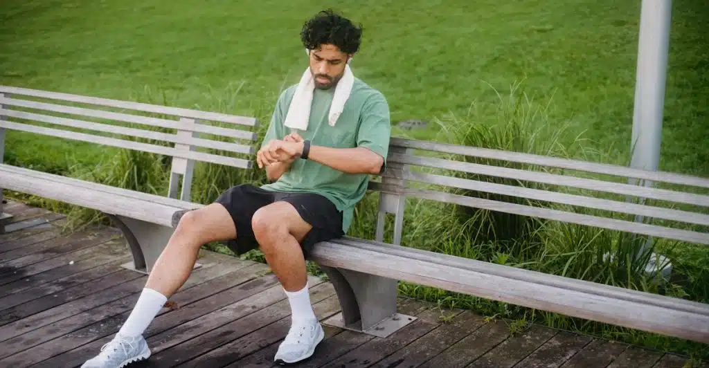 A man with a towel around his neck, sitting on a wooden bench outdoors, looking at his smartwatch on his left wrist.