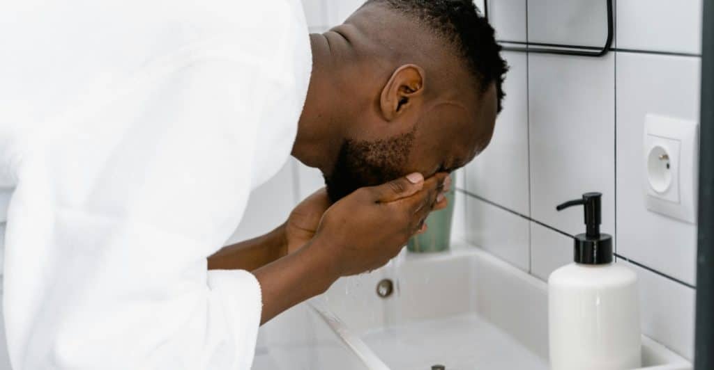 Man in a white bathrobe washing his face over a bathroom wash basin.