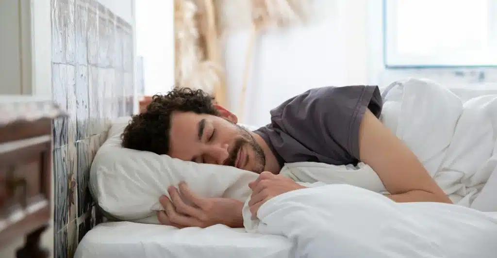 A man with curly hair sleeping on his side in a white bed with his hand near his face.