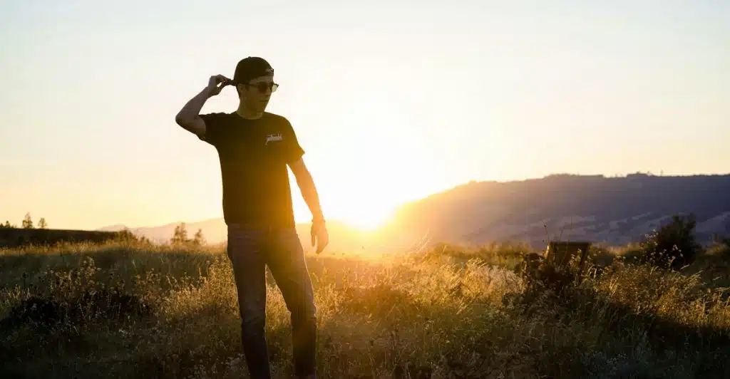 A man wearing a black t-shirt, sunglasses, and a baseball cap standing in a field of tall grass during sunset, with golden light illuminating the scene.
