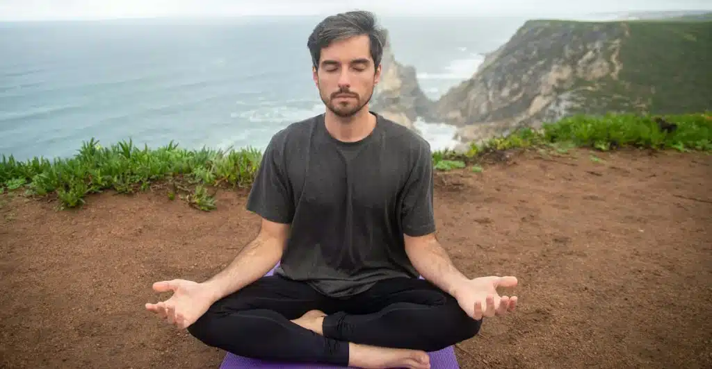 A man with dark hair and a beard sitting cross-legged on a purple yoga mat, meditating with his eyes closed on a cliff overlooking the ocean and rocky coastline.