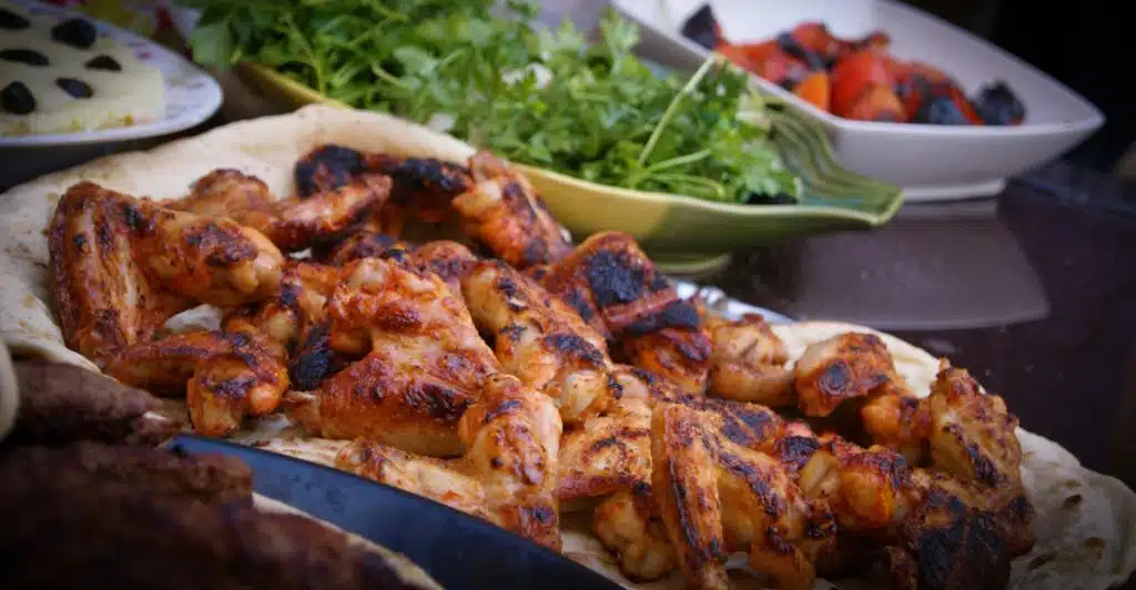 A platter of perfectly grilled chicken wings on flatbread, with bowls of fresh green herbs and roasted red vegetables in the background.