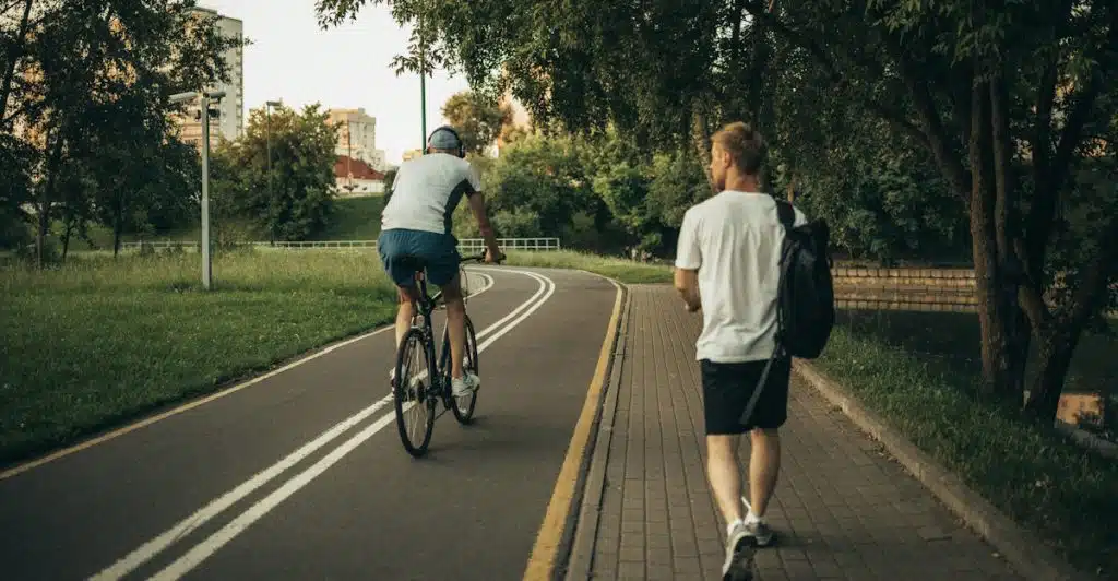 A man walking with a backpack on a paved path next to a grassy area, while another man rides a bicycle ahead of him on a parallel lane.