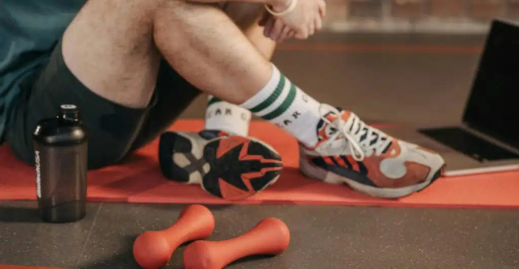 Two orange dumbbells on a red yoga mat with a person's legs and a laptop in the background.
