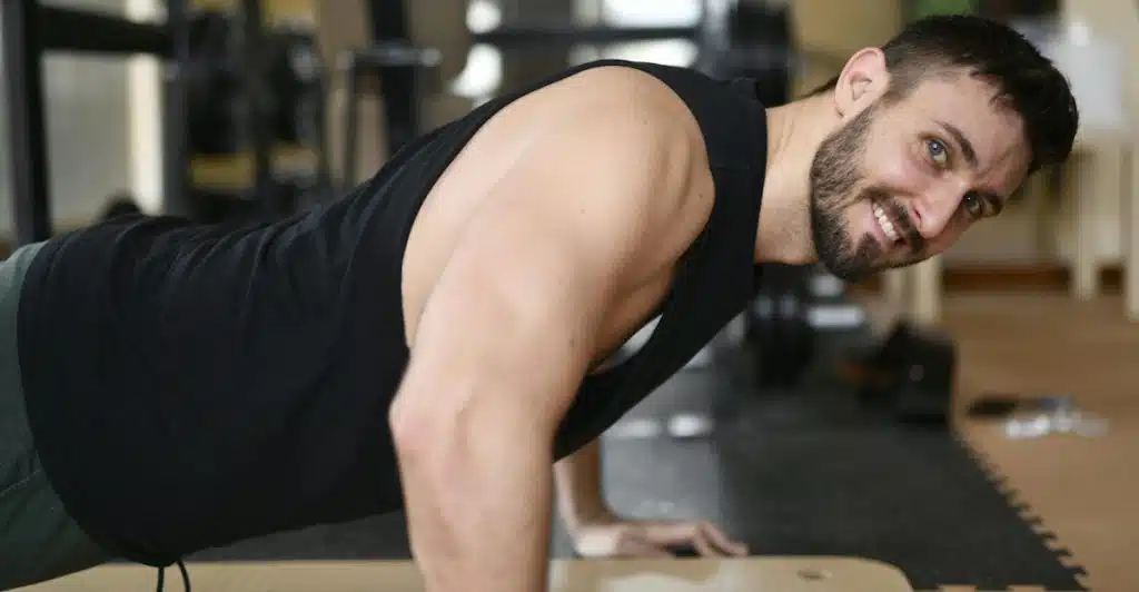 A man in a black tank top smiling while doing a push-up, looking towards the viewer.