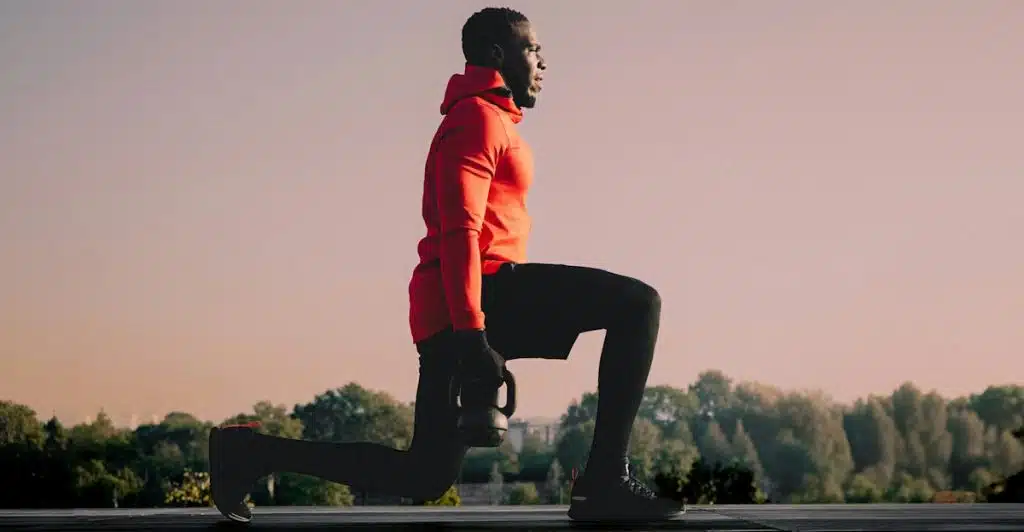A man in a red hoodie and black pants performing a lunge while holding a kettlebell outdoors.
