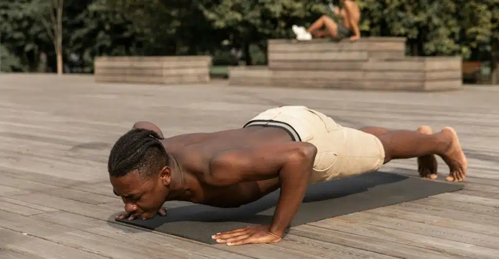 A shirtless man in beige shorts doing push-ups on a black yoga mat outdoors on a wooden deck.