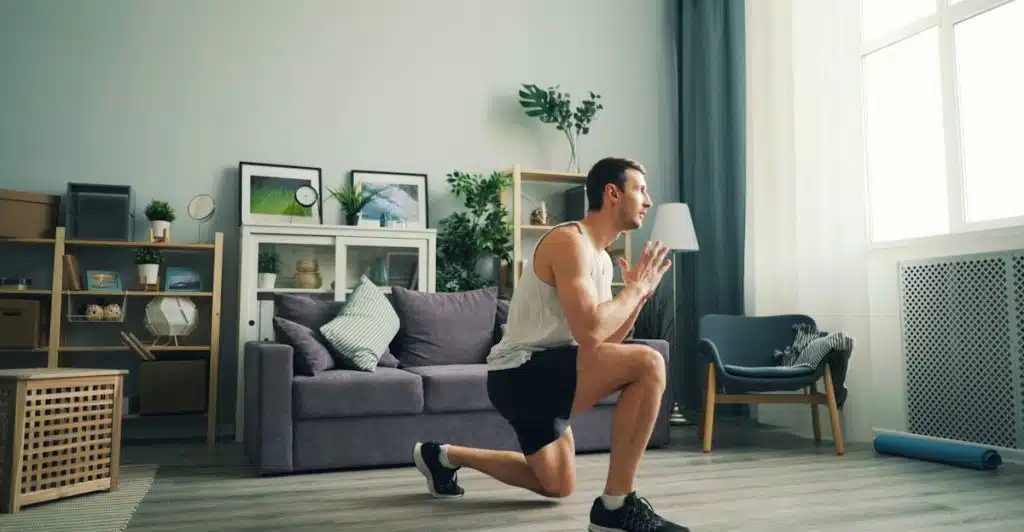 A man in a white tank top and black shorts doing lunges in a living room.