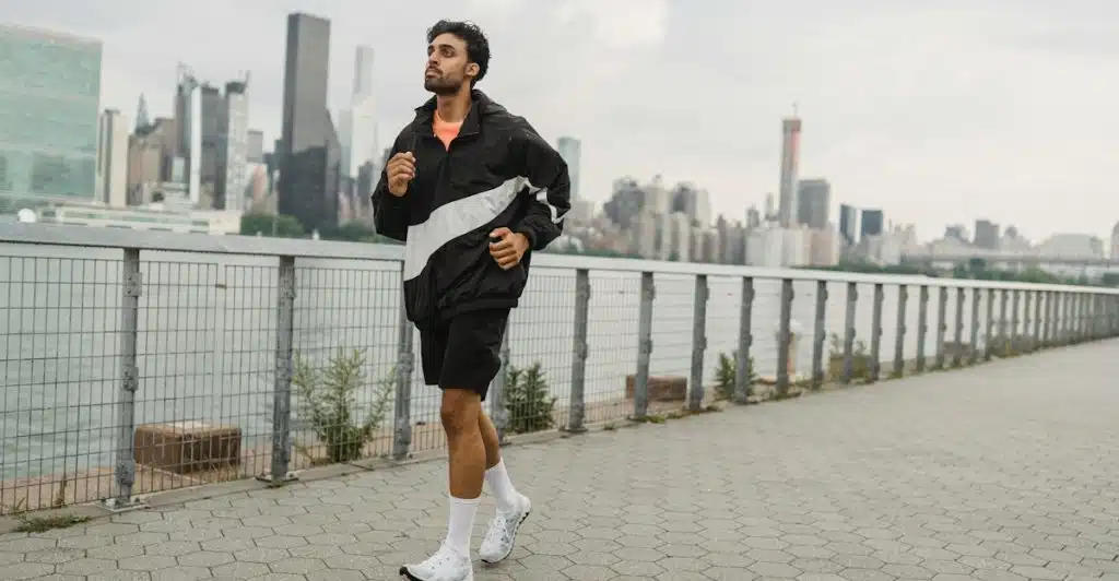 A man in a black and white jacket and black shorts jogging on a pathway with a city skyline in the background.