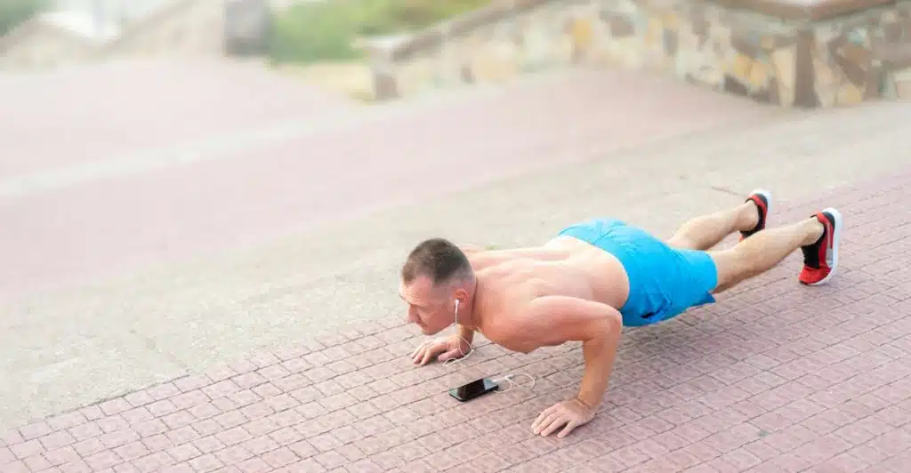 A shirtless man in blue shorts doing a plank on a paved surface with a phone and wired earphones in front of him.