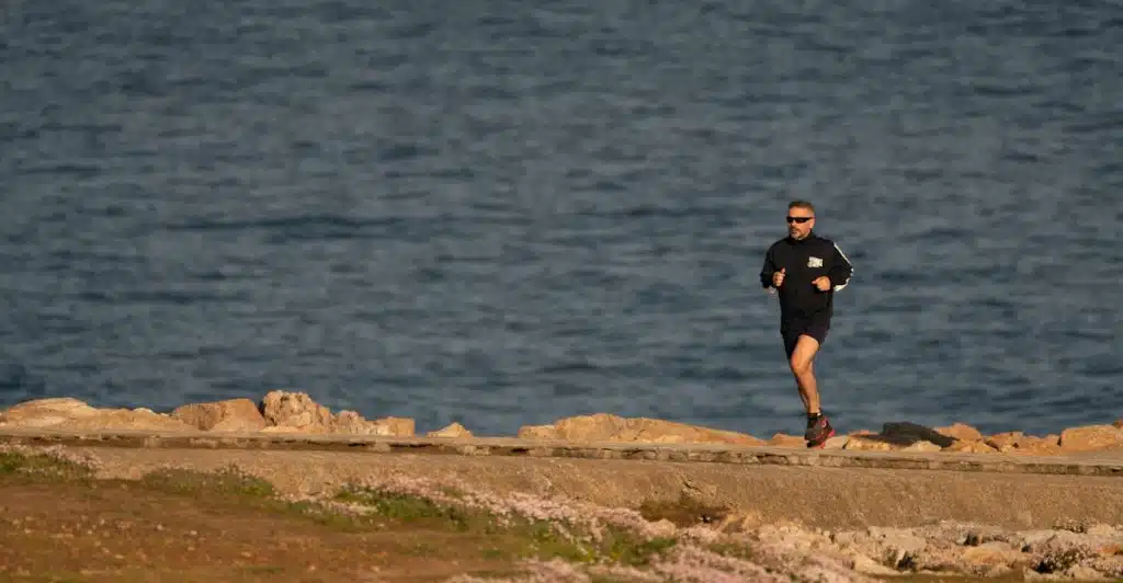 A man in sunglasses and a long-sleeved shirt running on a rocky path next to a body of water.