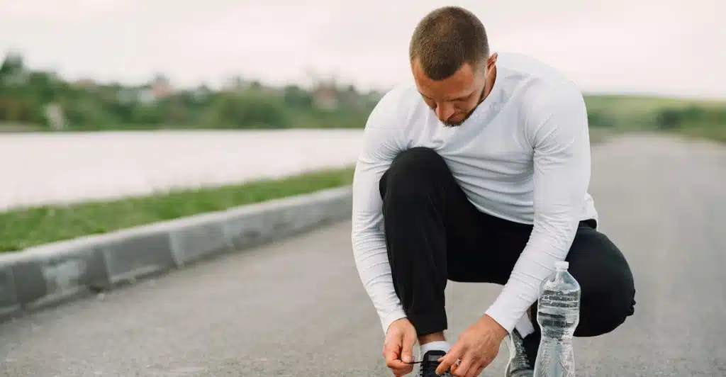 A Muscular man in white shirt and black pants tying his shoelaces on a road with a view of a lake in the background.