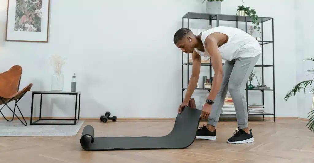 A man in a white tank top and gray sweatpants rolling up a grey yoga mat in a room with plants and furniture.