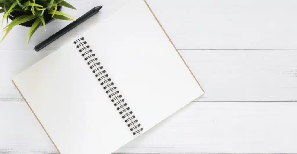 A blank spiral notebook with a black pen and a small green plant in a pot are arranged on a white wooden desk.