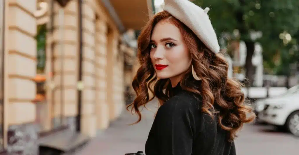 A woman with long wavy red hair wearing a black top and a white beret looks back over her shoulder while holding a coffee cup.